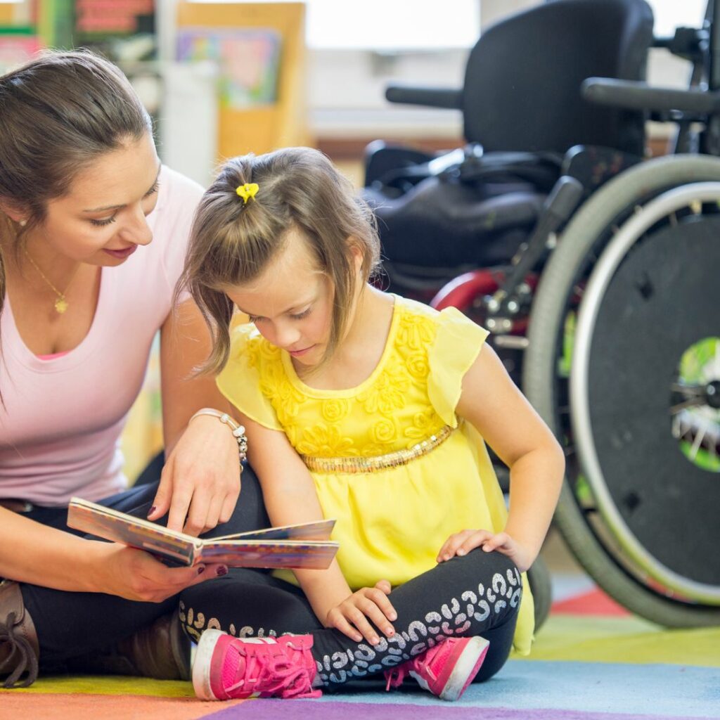 une femme portant un haut rose fait la lecture à une petite fille en situation de handicap, le fauteuil étant en fond d'image, la petite fille porte une robe jaune et des chaussures roses vifs.
