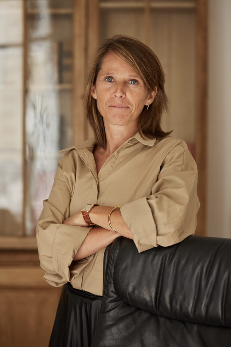 Maître Romy Collard avec une chemise beige et une jugpe marron devant son fauteuil dans son bureau ; dans le fond une bibliothèque