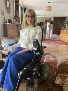 Inès Chometon, juriste du cabinet en situation de handicap, en fauteuil rouland, pantalon bleu et chemise blanche ; en arrière plan salle de séjour, tapis et parquet au sol, meuble dans le fond.