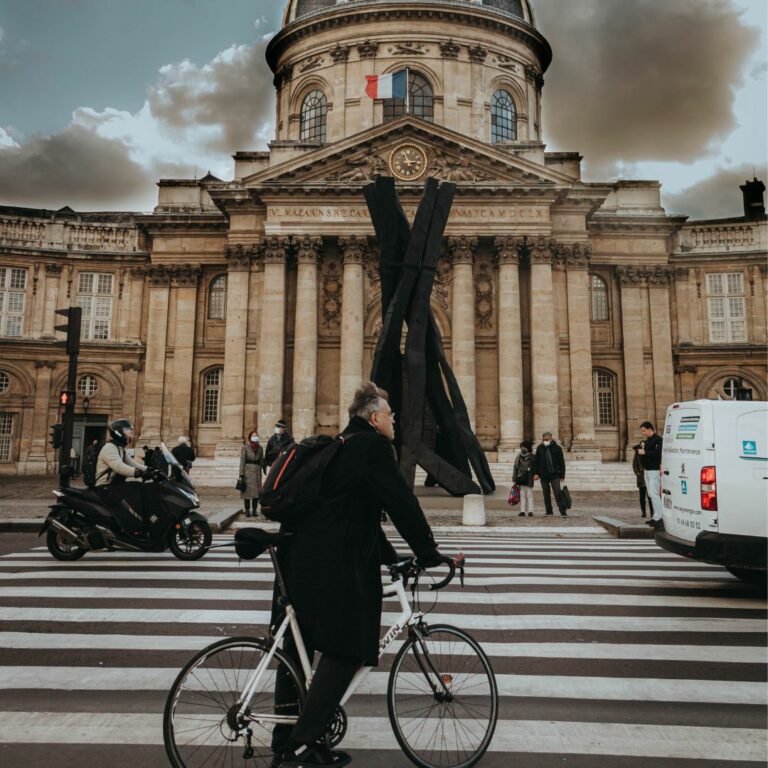 Homme à vélo sans casque en premier plan roulant sur les quais de Seine à Paris avec des voitures en fond et scooter. En arrière plan l'Académie française devant le pont des Arts.