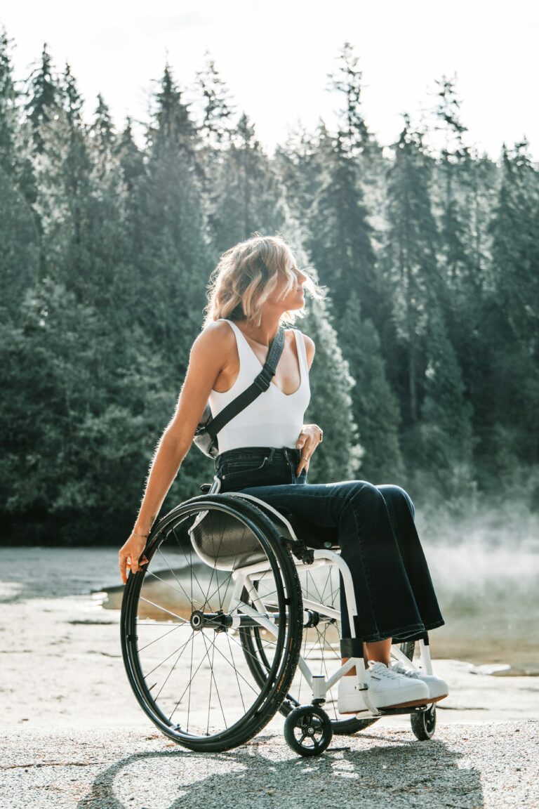 Jeune femme blonde sur un fauteuil roulant. Arrière plan la forêt et une belle luminosité, soleil. La jeune femme porte un débardeur blanc et un jean noir, et des baskets blanches