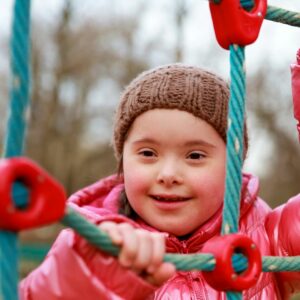 Une petite fille en situation de handicap, dans une erre de jeux ; elle porte une parka rouge et un bonnet marron, elle sourit.