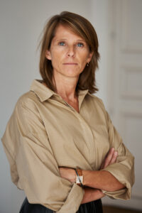 Maitre Romy Collard dans son bureau. Fond blanc. Elle prote une chemise beige et à les bras croisés devant.